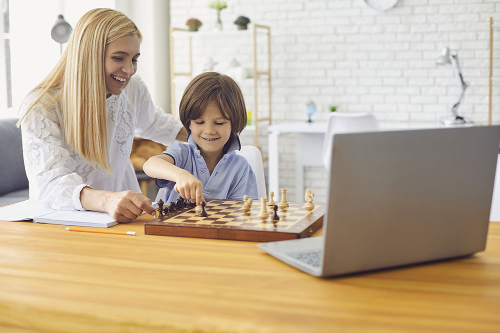 A mother plays chess with her young son while grandparents watch using Zoom.